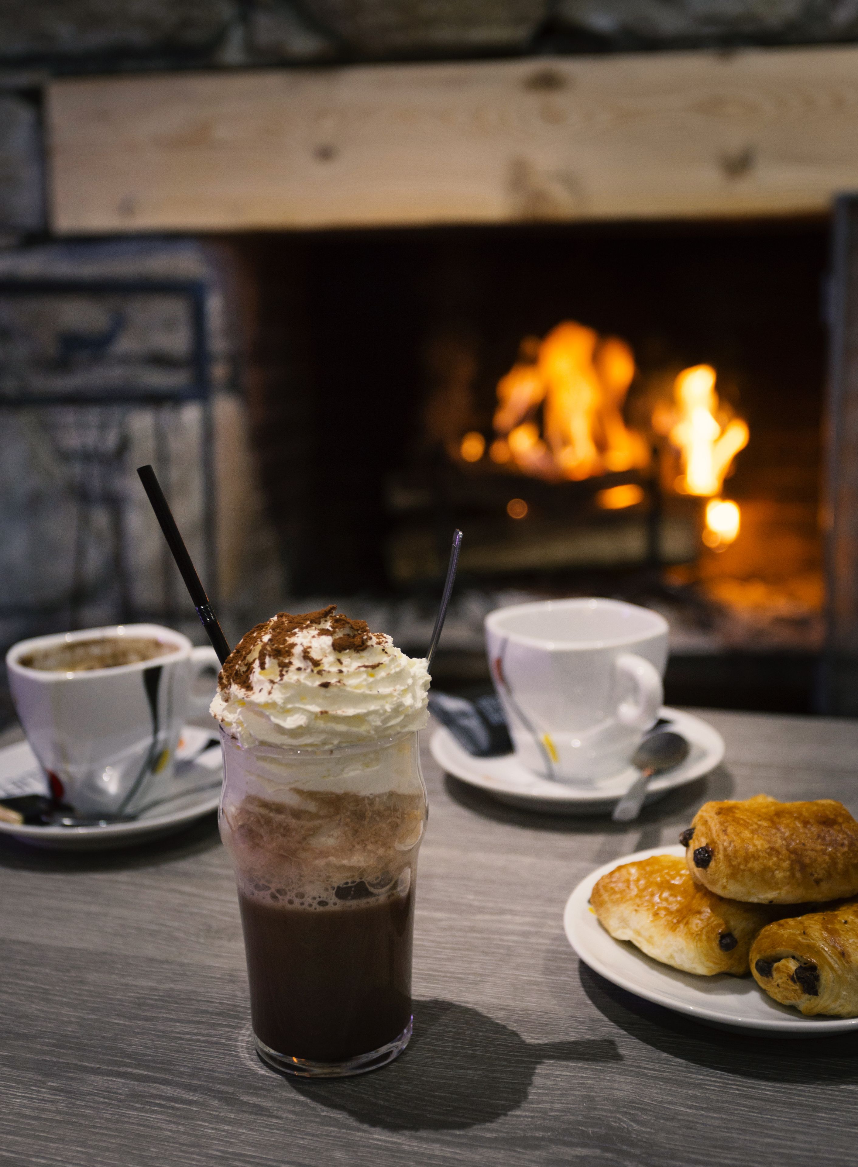 A table with a glass of layered beverage, two cups of coffee, and a plate of pastries. A fireplace with a fire is in the background.