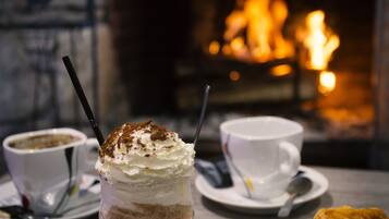 A table with a glass of layered beverage, two cups of coffee, and a plate of pastries. A fireplace with a fire is in the background.