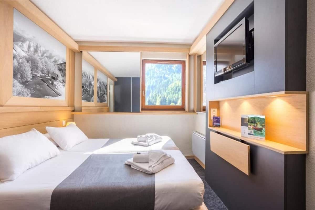 Ein Hotelzimmer mit Bett, Schreibtisch, Fernseher und einem Fenster mit Blick auf Bäume.