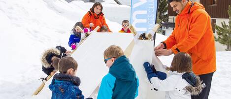 Un grupo de niños y adultos construyendo un muñeco de nieve en un entorno nevado al aire libre, con montañas al fondo.