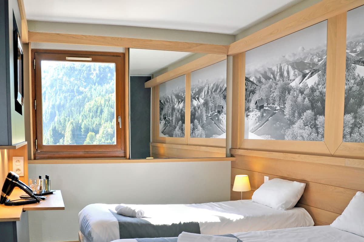 Ein Hotelzimmer mit zwei Betten, einem Schreibtisch und einem Fenster mit Blick auf Bäume und Berge.