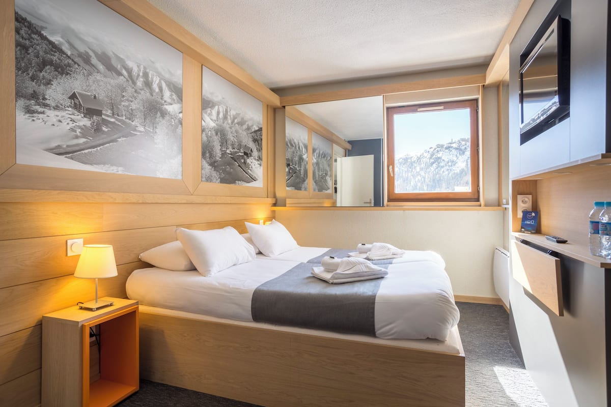 Ein Hotelzimmer mit Bett, Nachttisch mit Lampe, Spiegel und Fenster mit Bergblick.