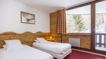 A room with two beds, a wooden headboard, a window with curtains, a radiator, and a framed picture on the wall.