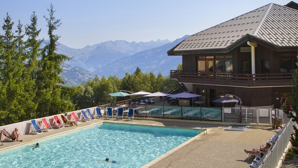 Un espace piscine avec une eau d’un bleu limpide, entouré de chaises longues et de parasols, et offrant une vue sur les montagnes en arrière-plan.