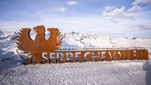Un letrero metálico con la palabra "Sierre Chevalier" y un águila estilizada, frente a un paisaje montañoso cubierto de nieve.