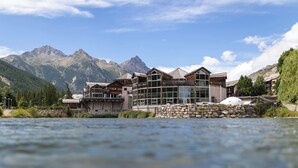 Un hotel moderno con fachada de vidrio, situado junto a un río y montañas al fondo.