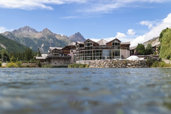 A modern hotel with a glass facade, located by a river with mountains in the background. - VVF Serre Chevalier Briançon (Monetier-les-Bains)