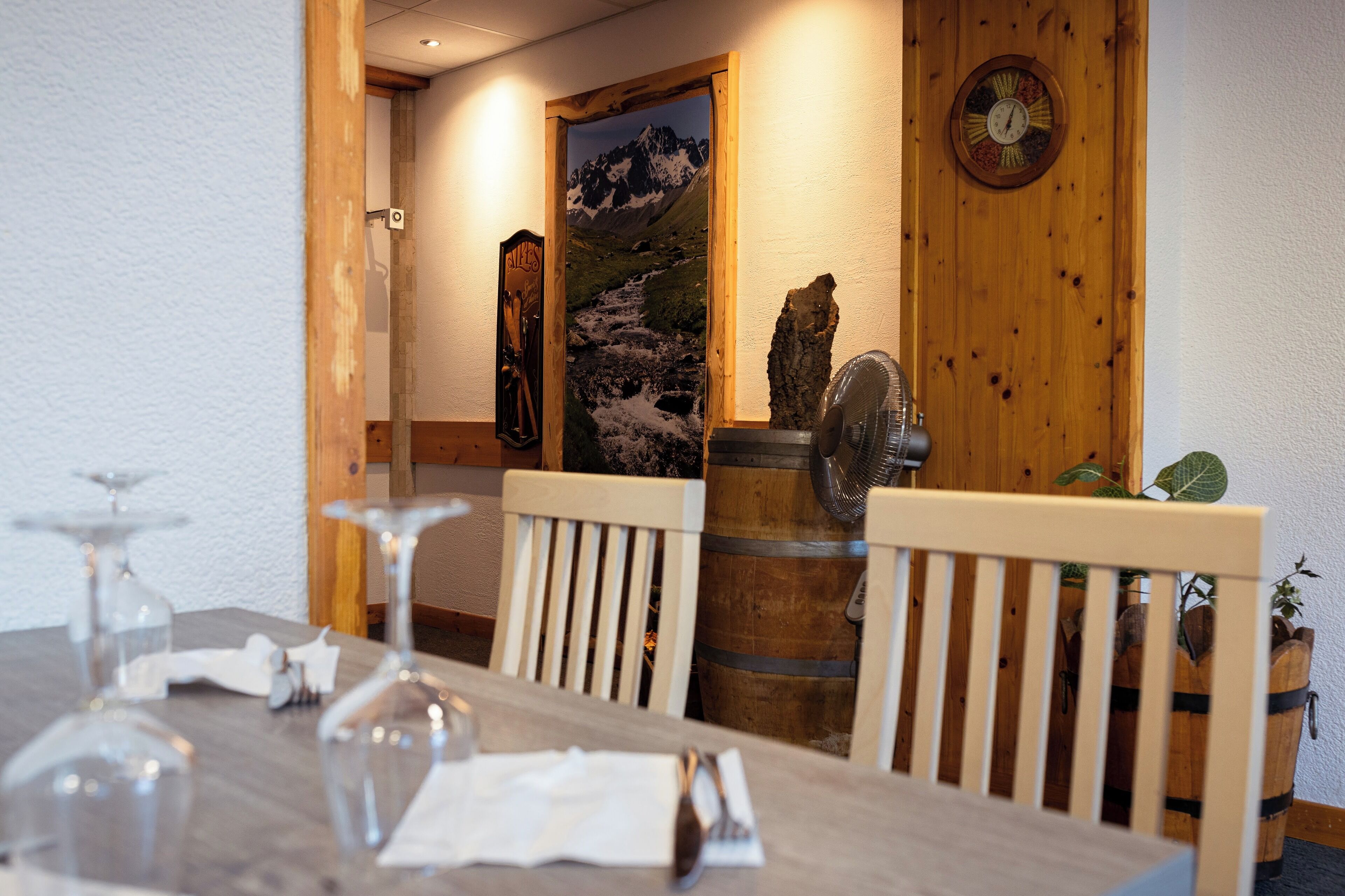 Un coin repas avec une table en bois, des chaises et un tonneau en bois. On y trouve une porte en bois ornée d’un tableau représentant un paysage de montagne, ainsi qu’une horloge au mur.