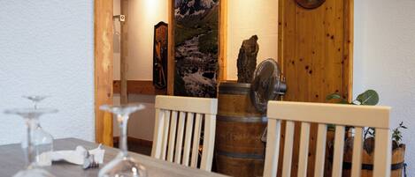 Un coin repas avec une table en bois, des chaises et un tonneau en bois. On y trouve une porte en bois ornée d’un tableau représentant un paysage de montagne, ainsi qu’une horloge au mur.
