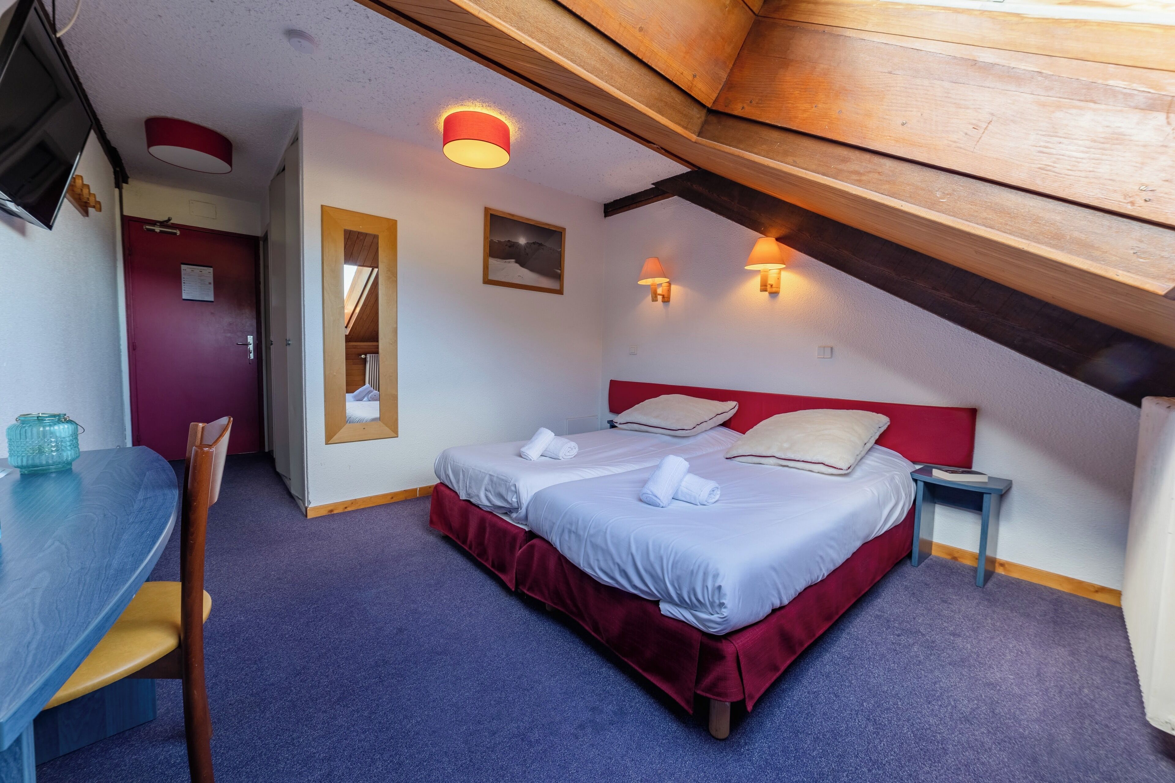 A bedroom with a bed, a desk, and a chair. There is a red door and a mirror on the wall.