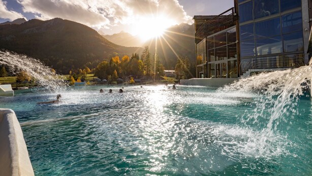 Una piscina con una cascada, rodeada de montañas y un edificio con grandes ventanales.