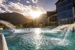 A swimming pool with a waterfall feature, surrounded by mountains and a building with large windows. - VVF Serre Chevalier Briançon (Monetier-les-Bains)