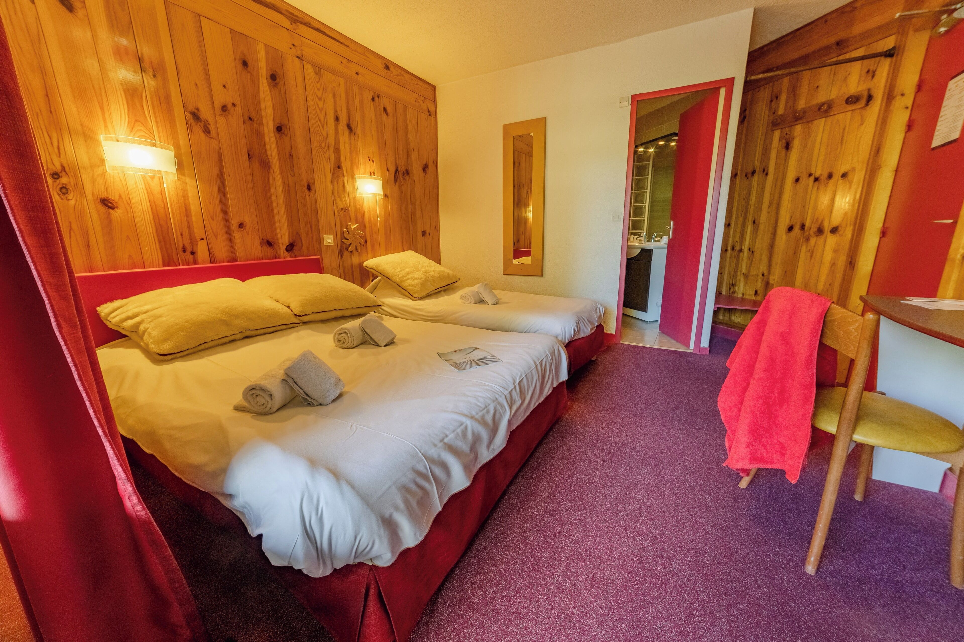 A hotel room with two beds, wooden paneling, a red curtain, a small table, and a chair.
