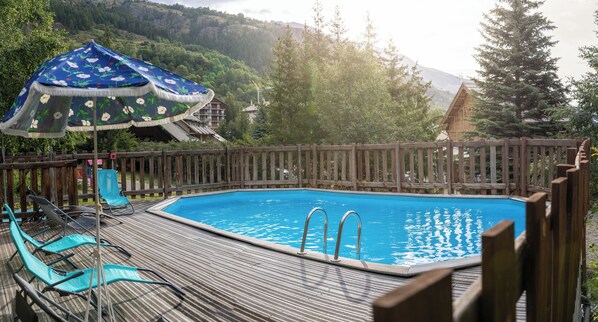 A wooden deck with a swimming pool, lounge chairs, and a decorative umbrella. - VVF Serre Chevalier Briançon (Monetier-les-Bains)