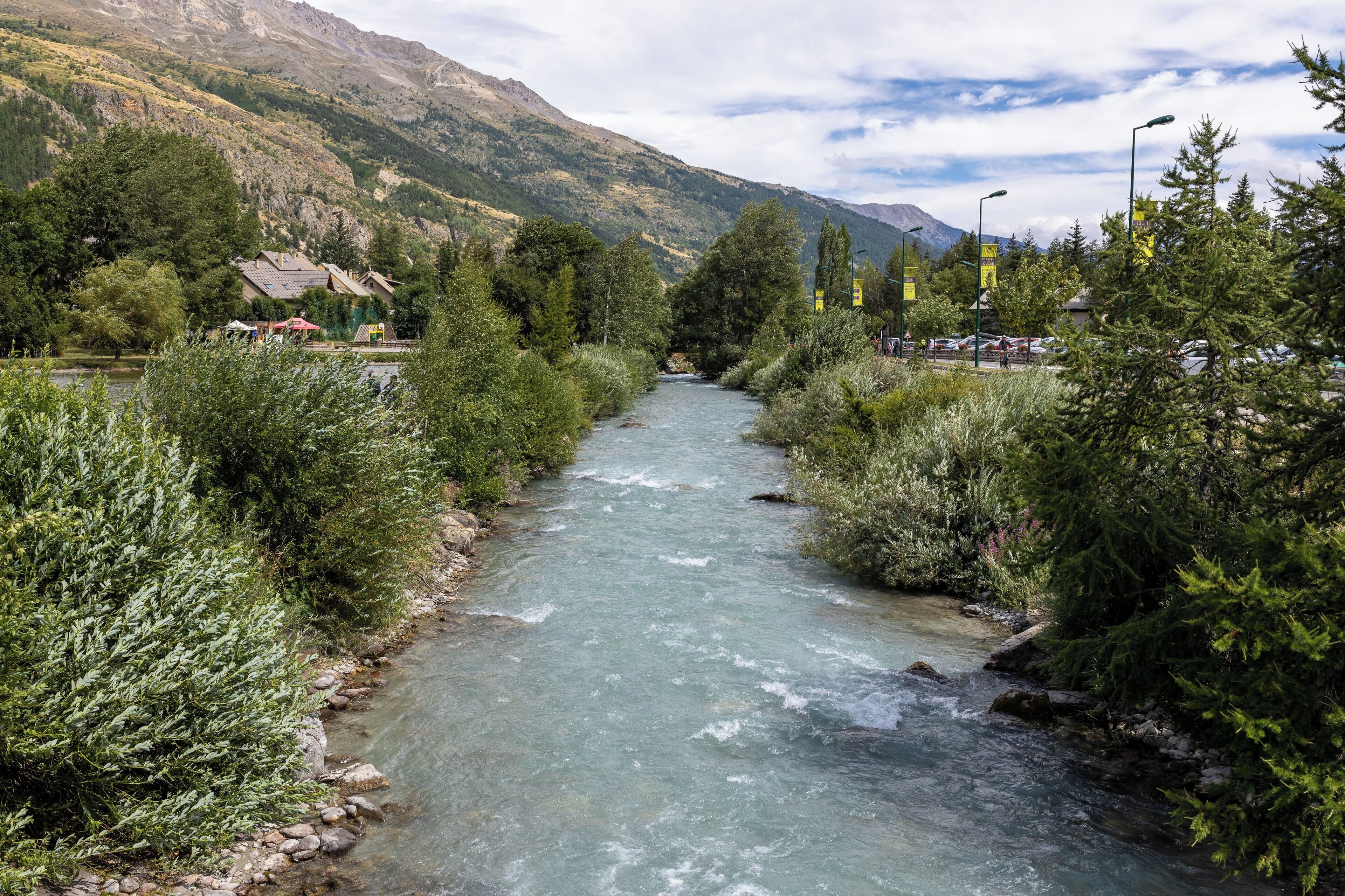 A river flowing through a valley with mountains in the background and a small settlement with houses and a road nearby.