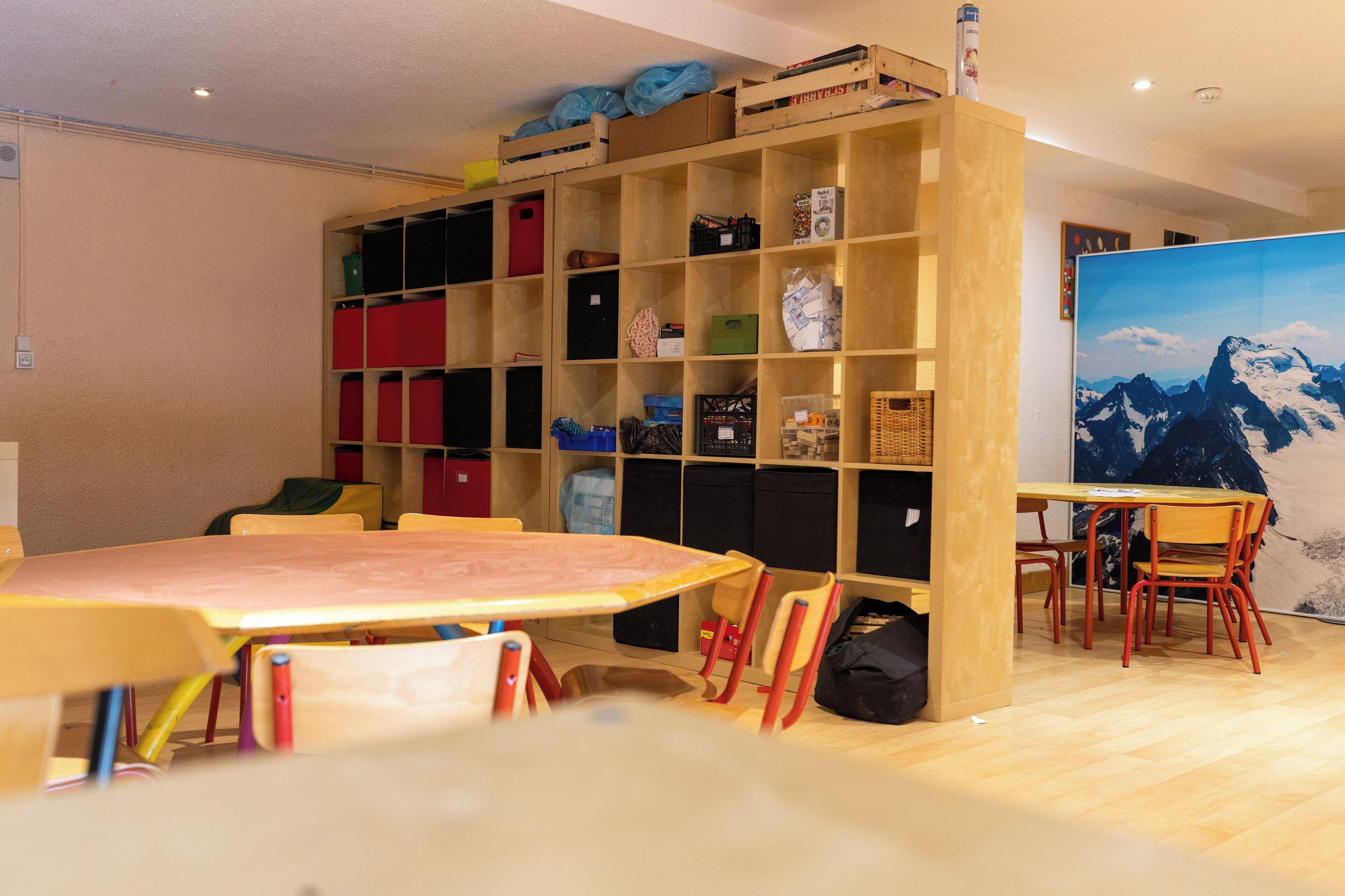 A room with a wooden bookshelf, a table with chairs, and a painting of mountains.