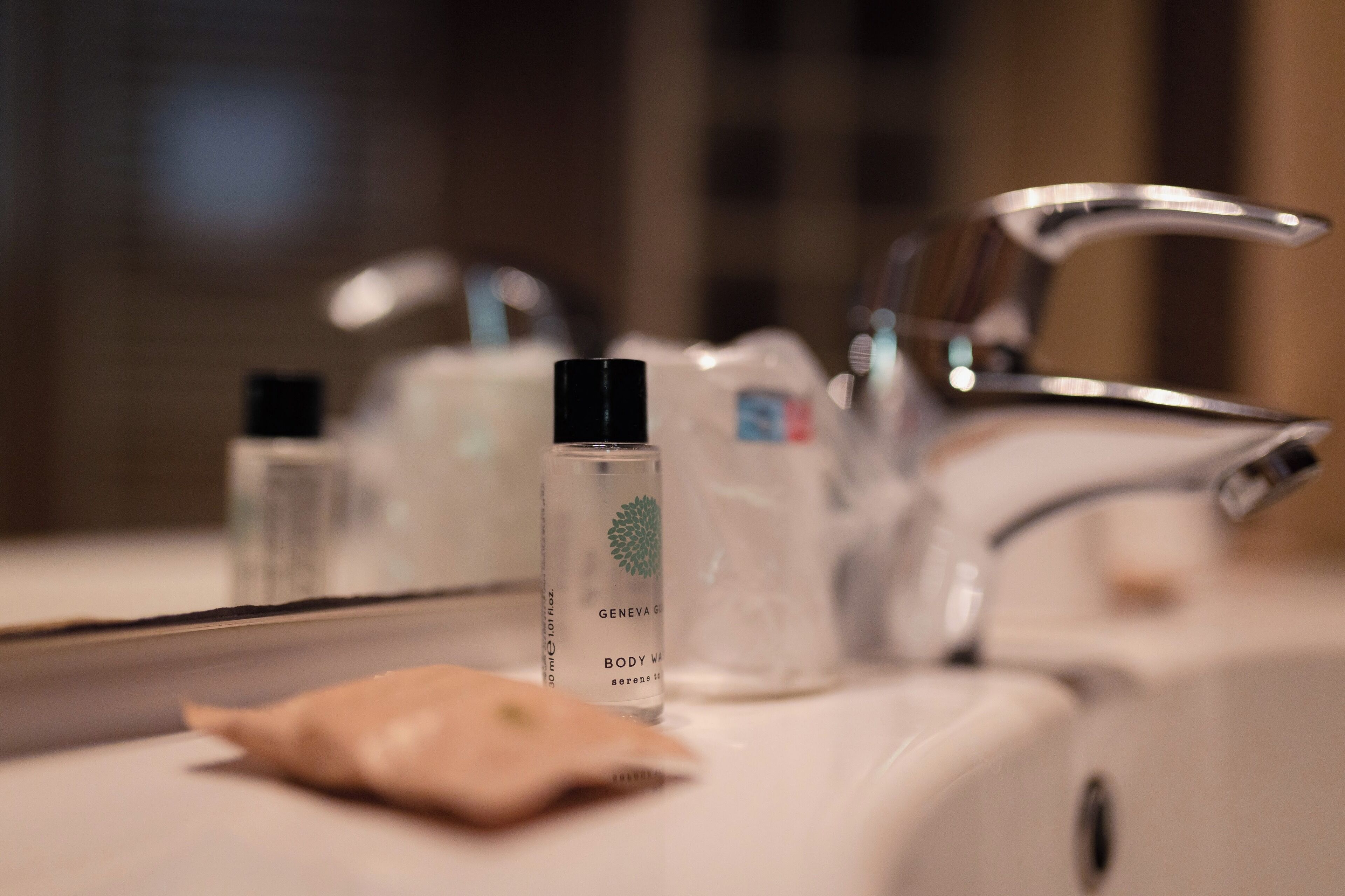 Une salle de bain équipée d’un lavabo, d’un miroir et de produits de toilette, dont un flacon étiqueté « GEL DOUCHE GENÈVE ».