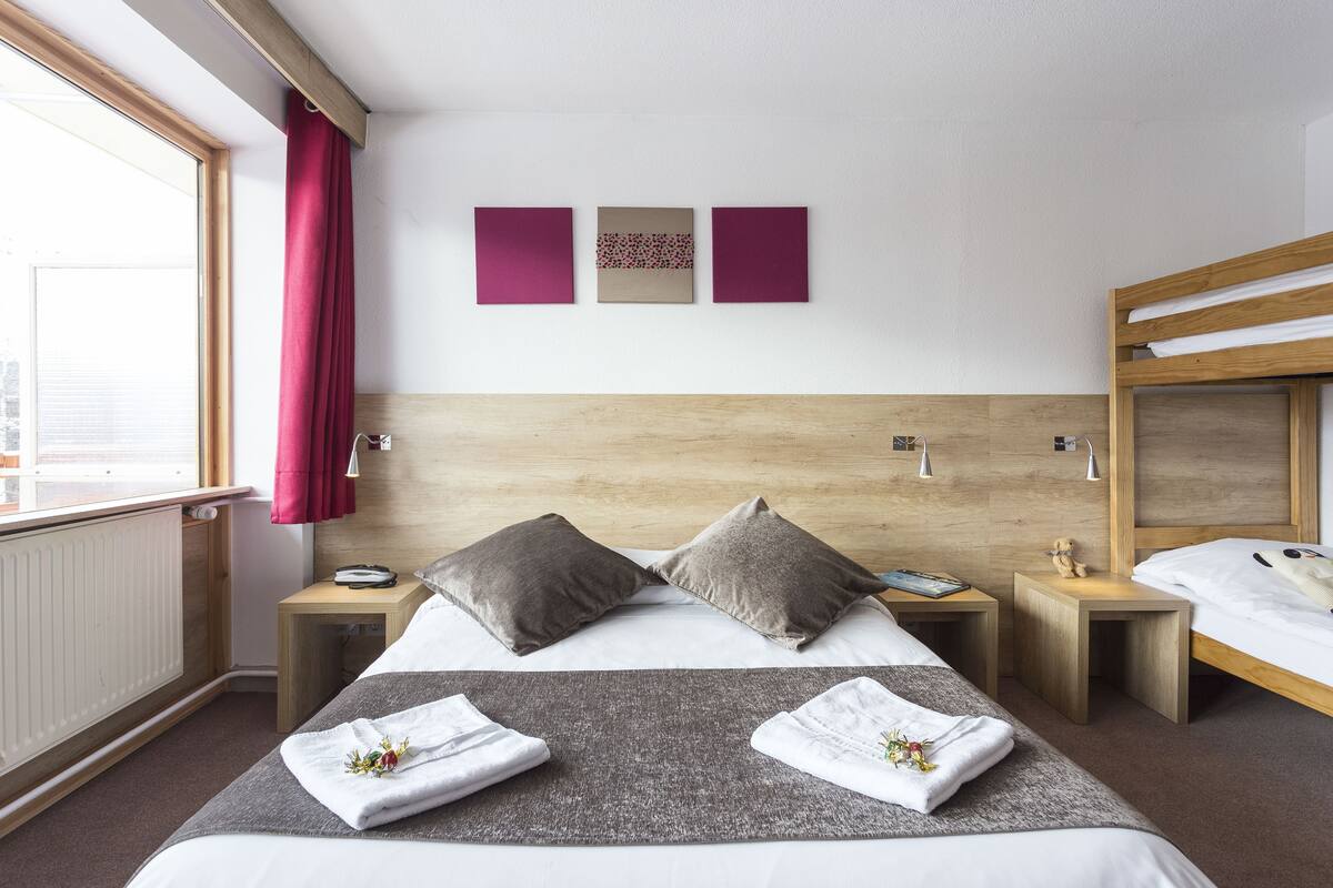 A hotel room with a bed, two bedside tables, a wooden headboard, a window with a view, and three wall-mounted artworks.