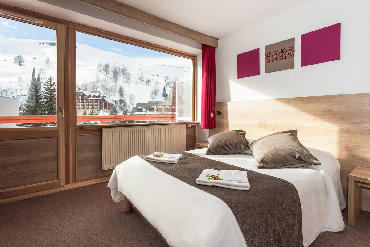 A hotel room with a large window, wooden headboard, two beds, and a view of a snowy mountain.
