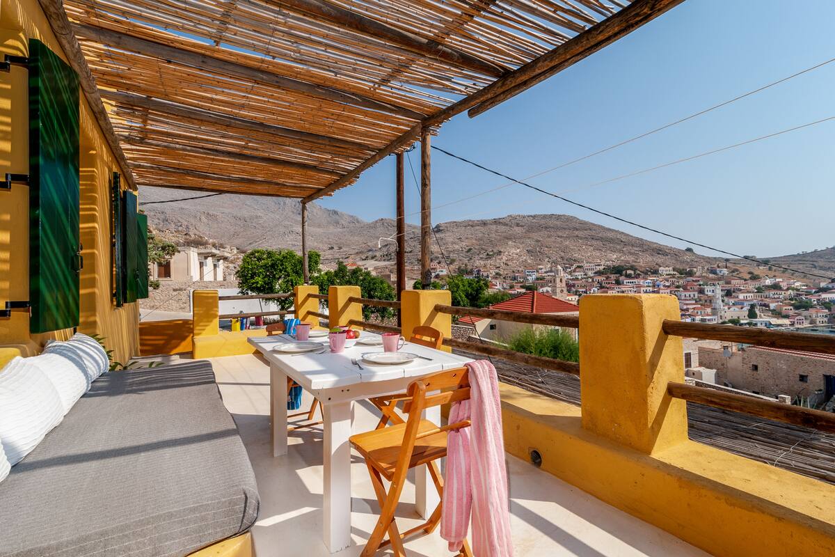 Um terraço com mesa e cadeiras brancas, uma pérgula de madeira e vista para uma cidade e montanhas ao fundo.