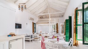 A bright, open-plan living and dining area with white furniture, a ceiling fan, and a TV mounted on the wall. - Atlantis Houses (Halki)