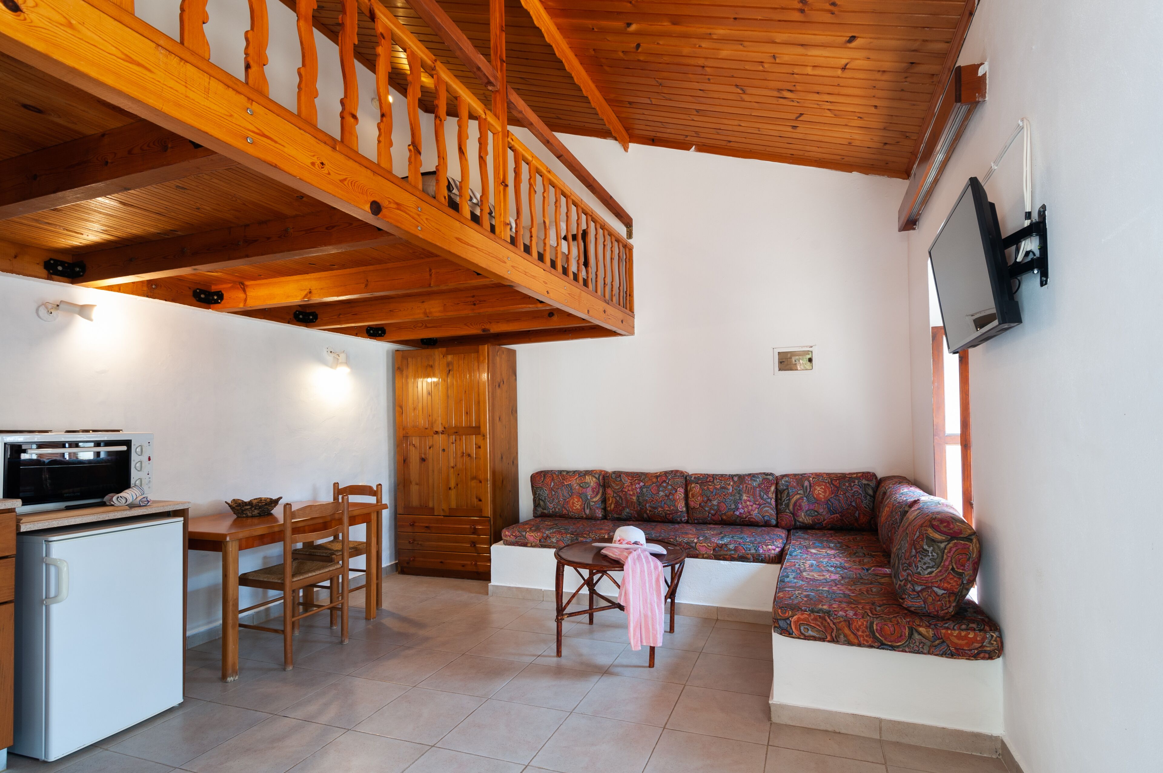 A cozy living area with a wooden staircase, a sofa, a dining table with chairs, a small refrigerator, and a television mounted on the wall.