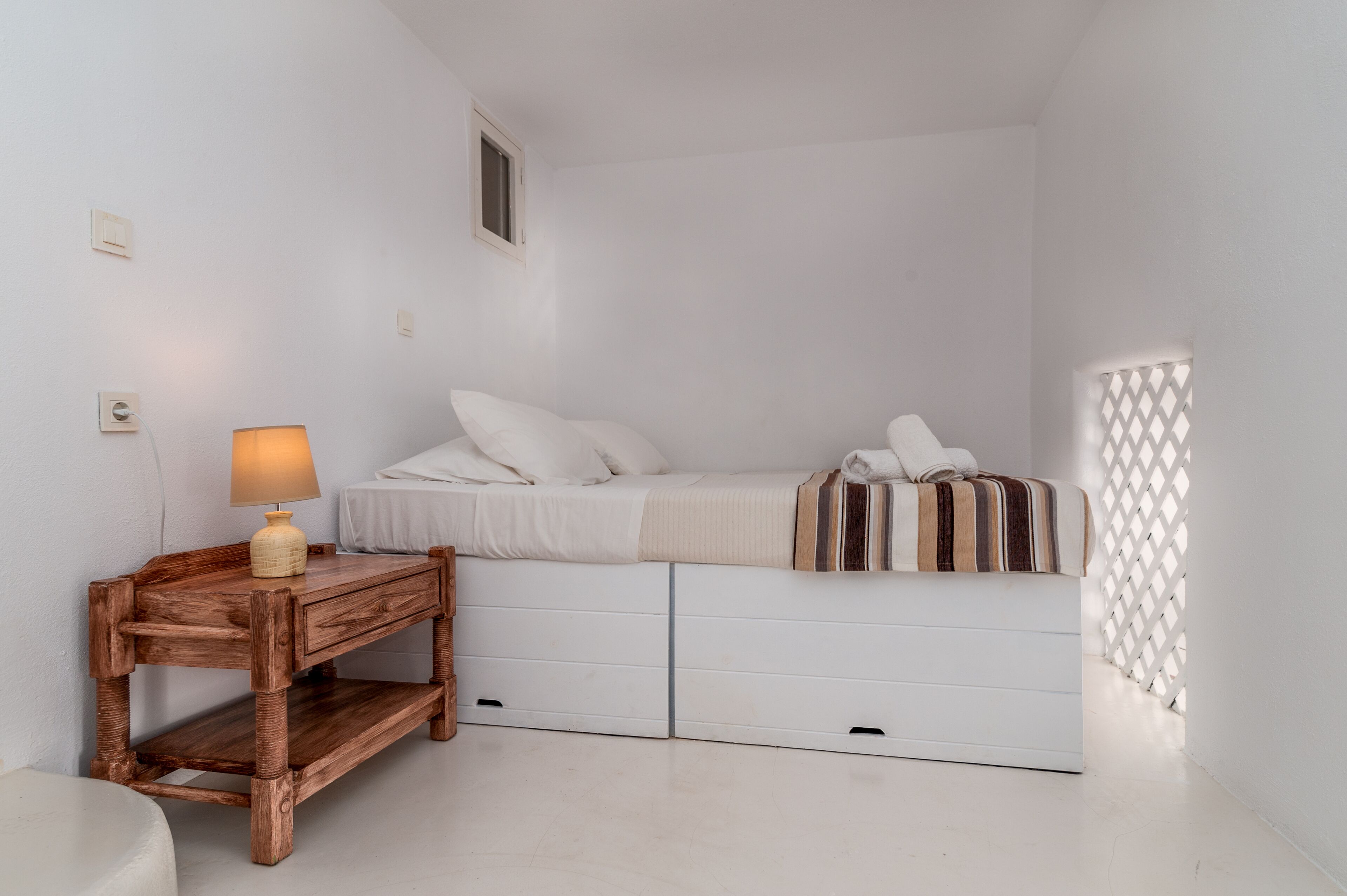 A small, white bedroom with a single bed, a wooden nightstand with a lamp, and a white wardrobe.