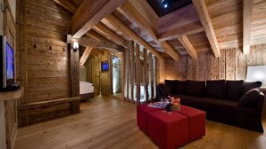 Ein rustikaler Innenraum einer Holzhütte mit Holzboden, einem roten Pouf, einem dunklen Sofa und einem Fernseher in der Ecke.