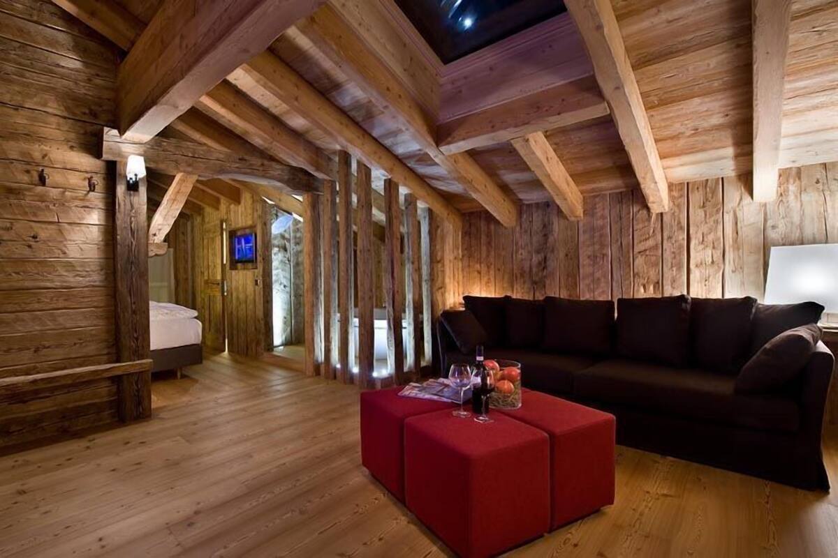 Interieur van een blokhut met een houten vloer, een rode poef, een donkere bank en een televisie in de hoek.