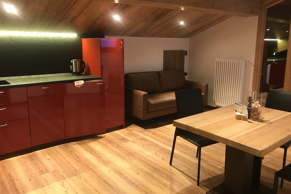 Een compacte keuken met een rode koelkast, een houten eettafel en een bruine bank.