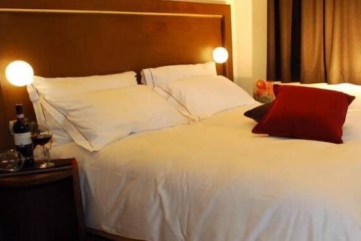 Een net opgemaakt bed met witte lakens en een rood kussen, een houten nachtkastje met een fles en glazen, en een rode stoel in een hotelkamer.