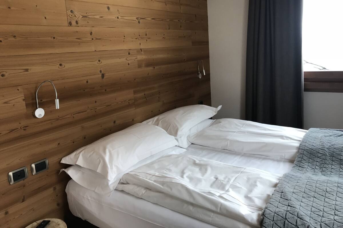Een kamer met houten lambrisering, een bed, wit beddengoed en een raam met gordijn.