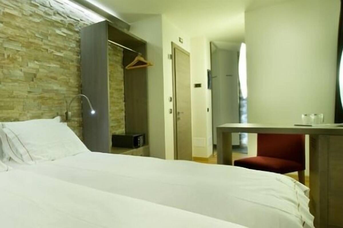 Een hotelkamer met een stenen muur, een bed met witte lakens, een bureau met stoel en een spiegel.