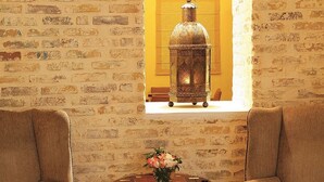 Una habitación con pared de ladrillo, dos sillones, una mesita con un florero y una ventana con un farol.