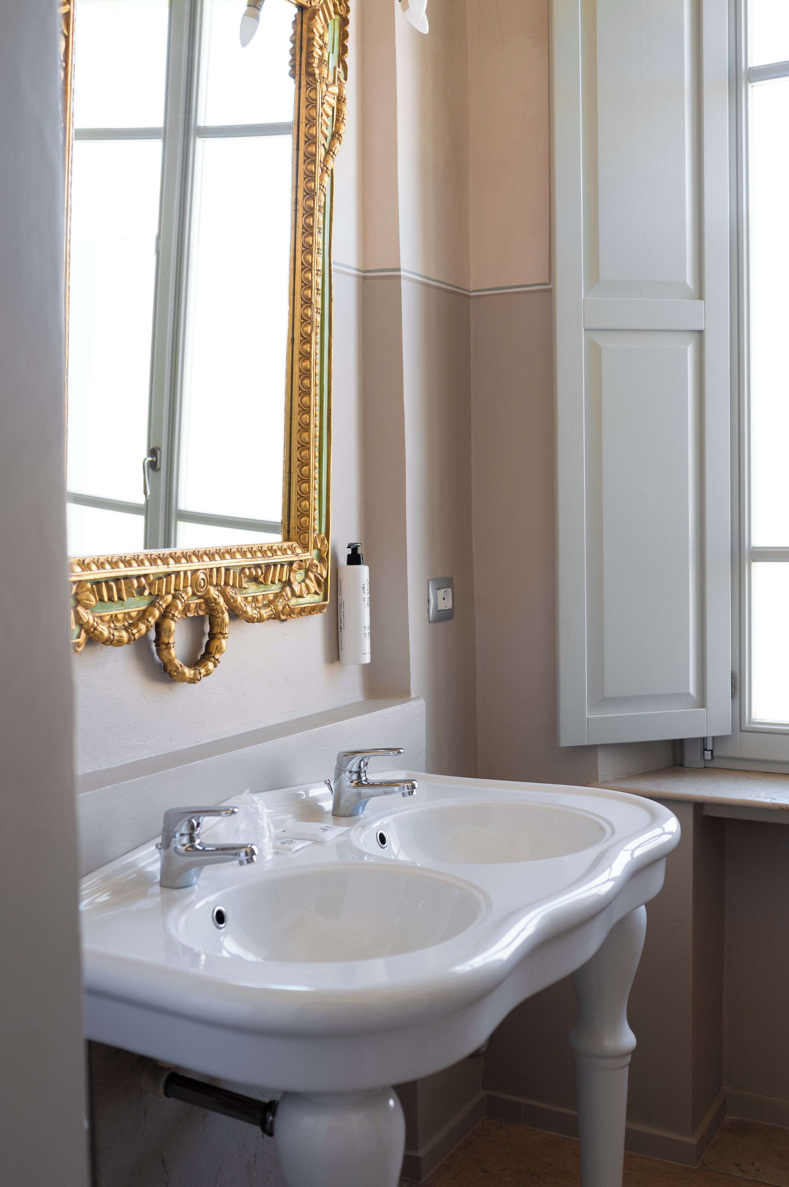 A bathroom with a double sink, a gold-framed mirror, and a window with a view.