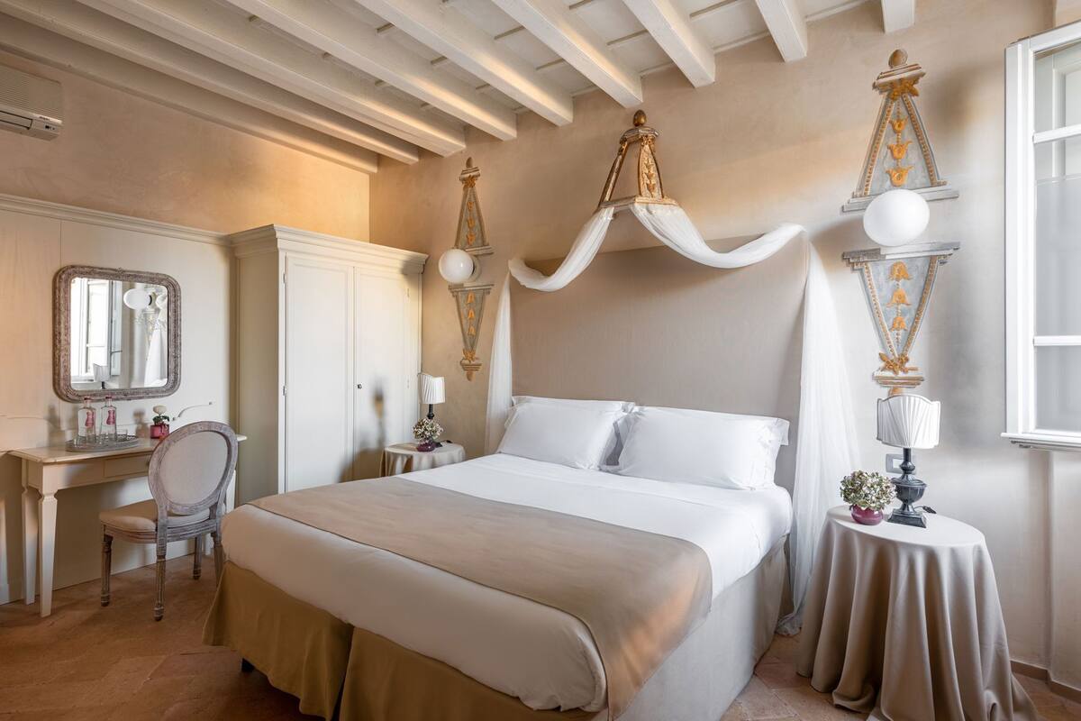 A bedroom with a bed, a vanity with a mirror, a chair, and decorative wall sconces.
