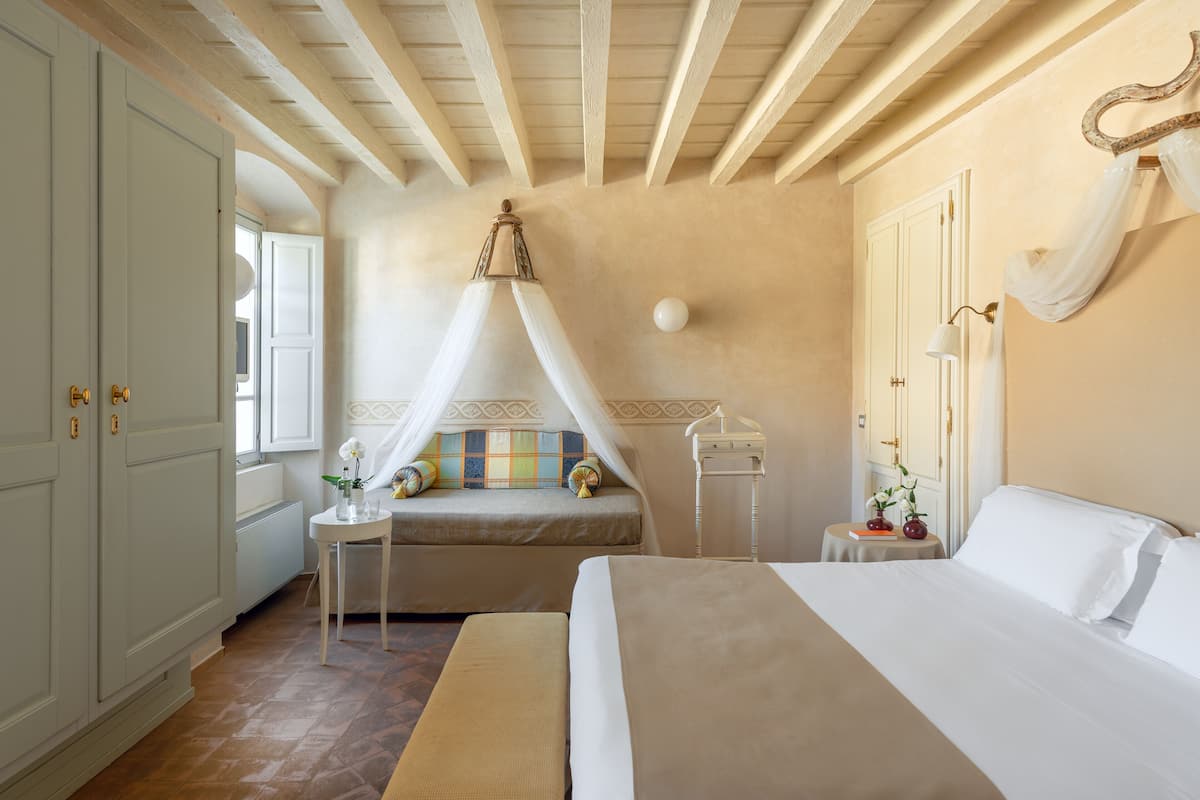 A bedroom with a large bed, a bedside table, a window with curtains, a bedside lamp, and a wall-mounted hook.
