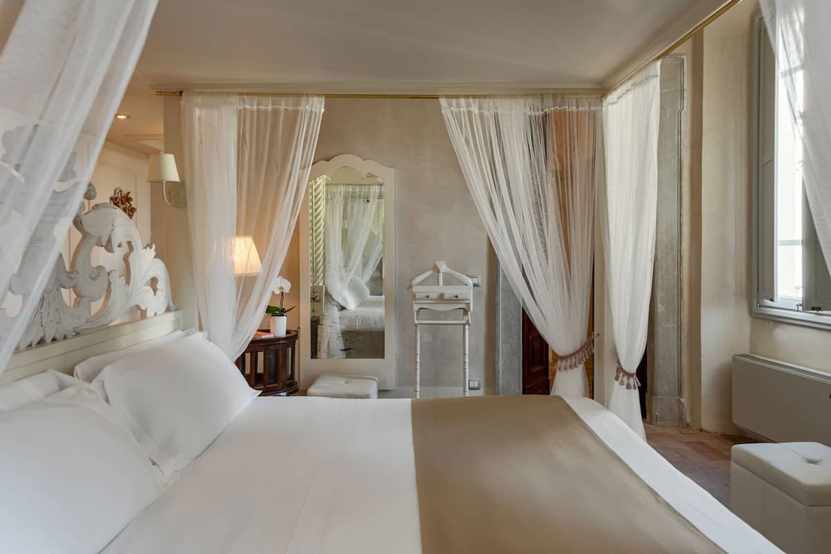 A large bed with white linens, a bedside table with a lamp, and a mirror with a decorative frame.
