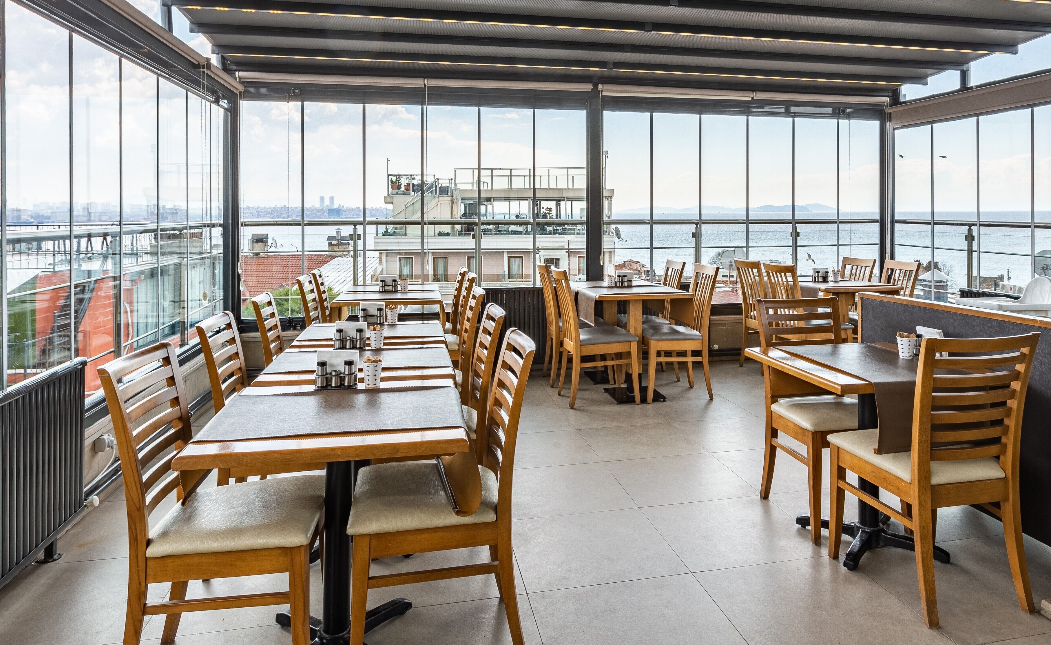Un espacio de comedor al aire libre con mesas y sillas de madera, amplias ventanas que ofrecen vistas a la ciudad y al agua.