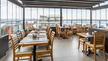 Un espacio de comedor al aire libre con mesas y sillas de madera, amplias ventanas que ofrecen vistas a la ciudad y al agua.