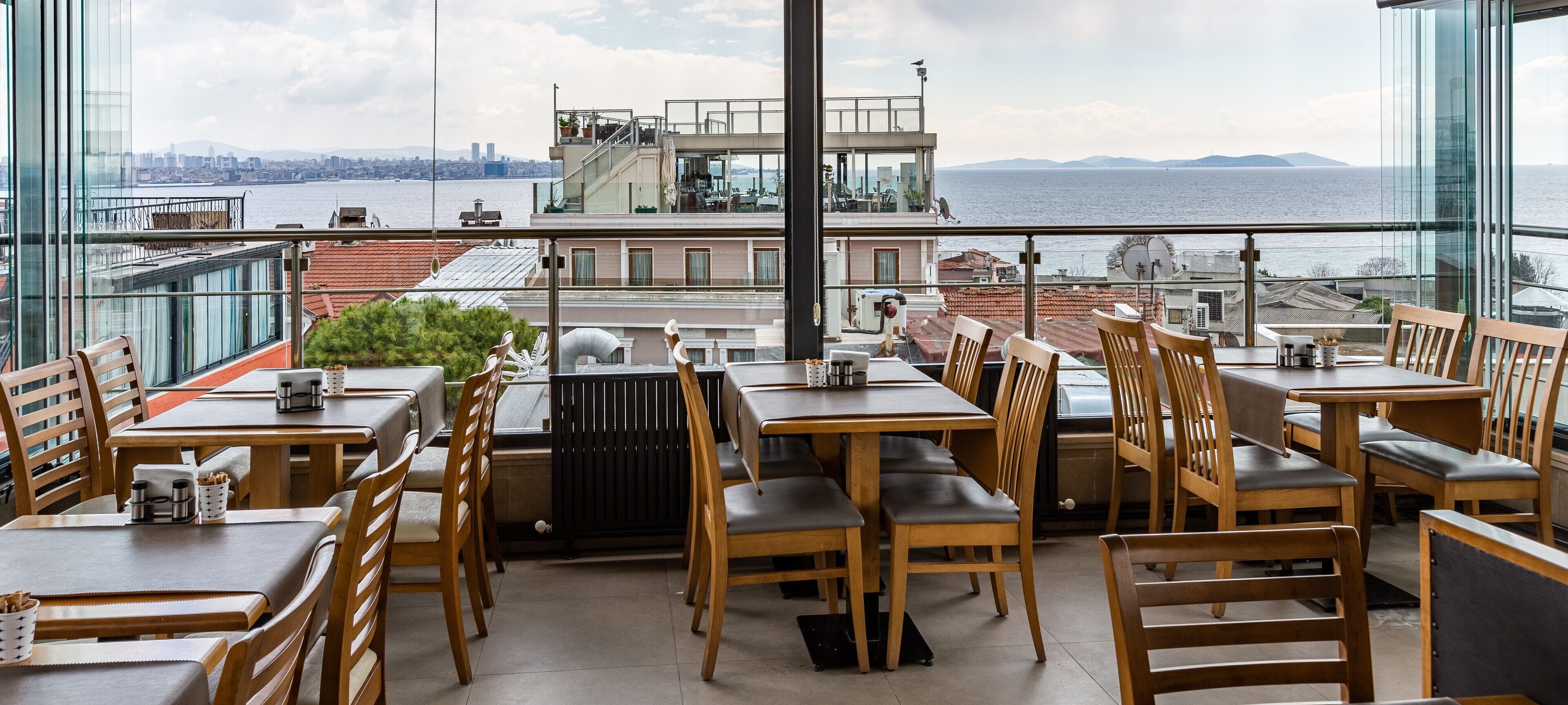 A restaurant with wooden tables and chairs, large windows offering a view of the sea and city skyline.