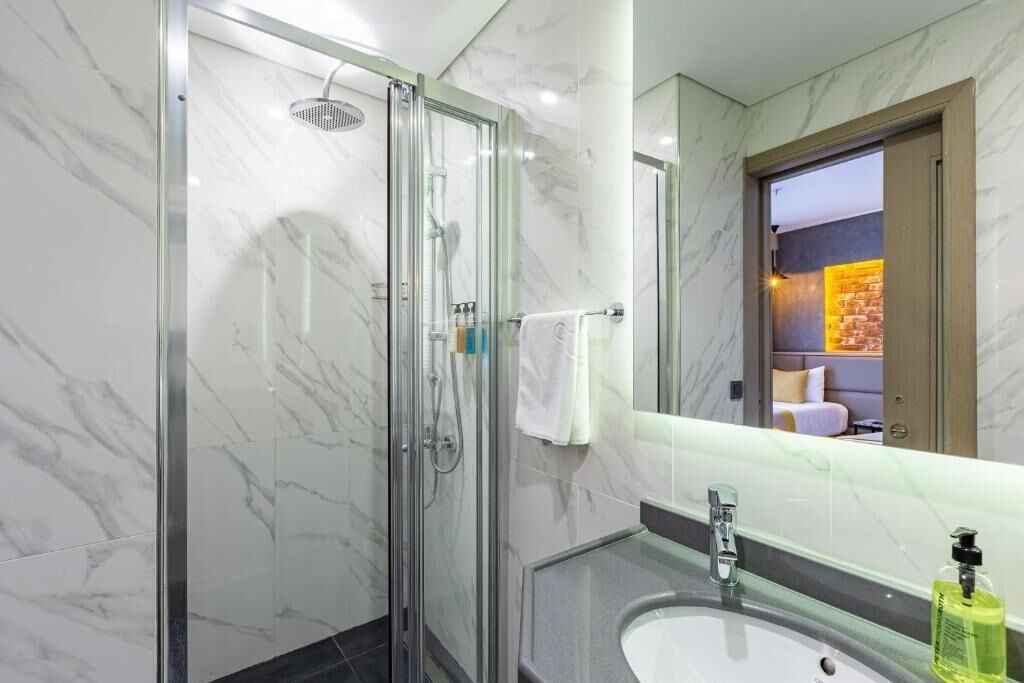 A modern bathroom with a glass-enclosed shower, a sink with a mirror, and a view of a room with a brick wall and a bed.