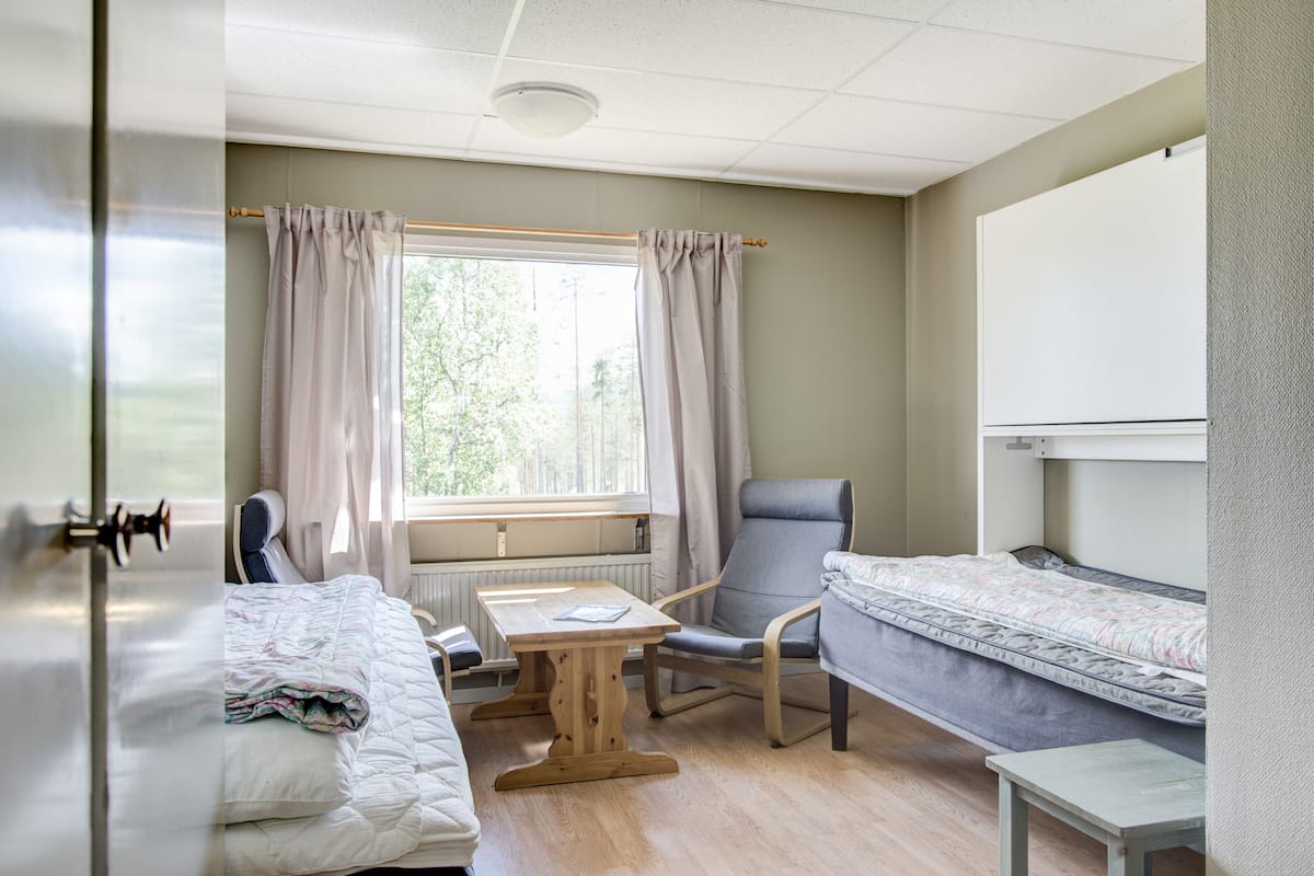 Ein Zimmer mit einem Bett, einem Sessel, einem Tisch und einem Fenster mit Vorhängen.