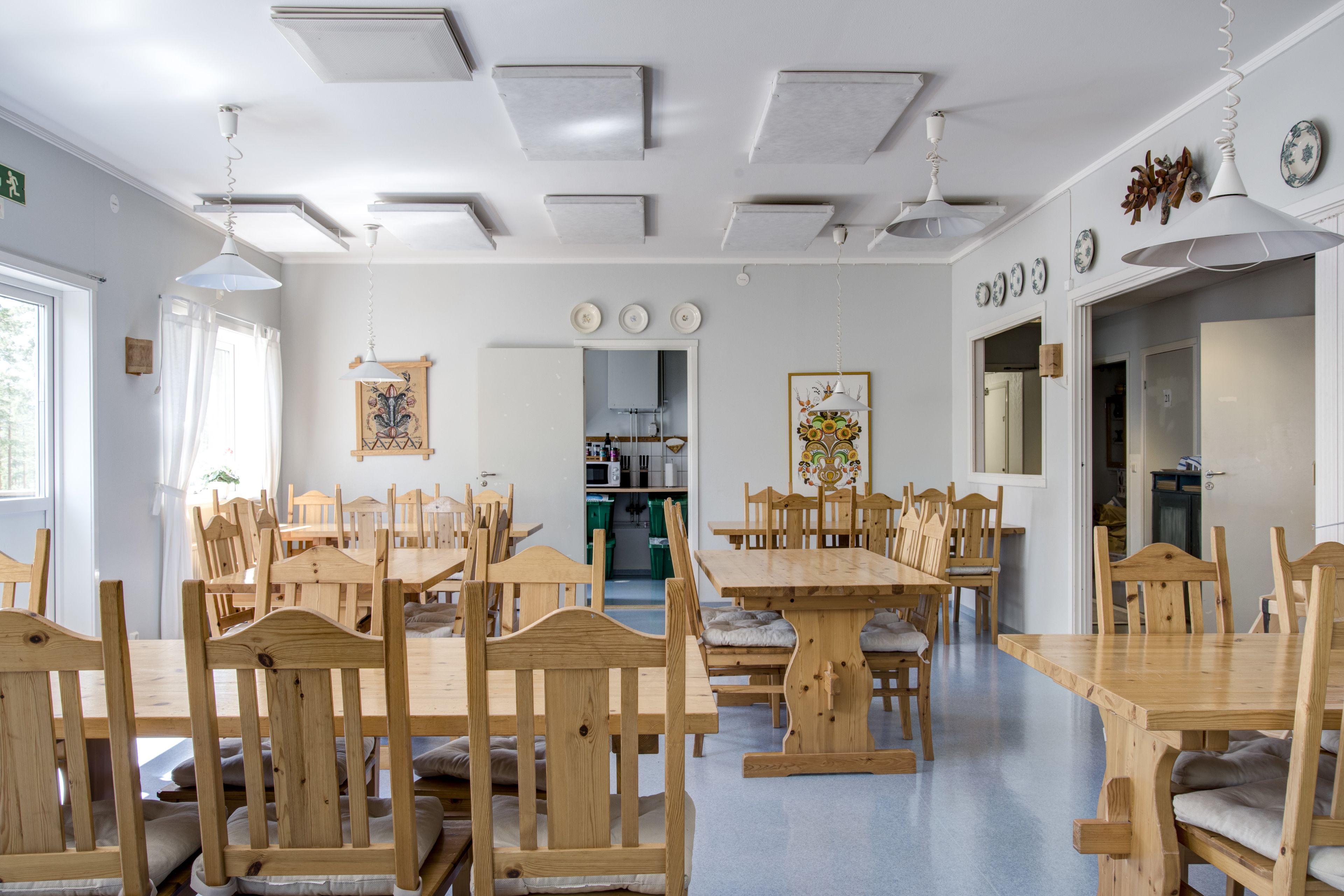 Un comedor con mesas y sillas de madera, una cocina al fondo e iluminación empotrada en el techo.