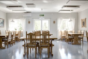 Uma área de refeições com mesas e cadeiras de madeira, janelas amplas com cortinas e um sinal de saída no teto.