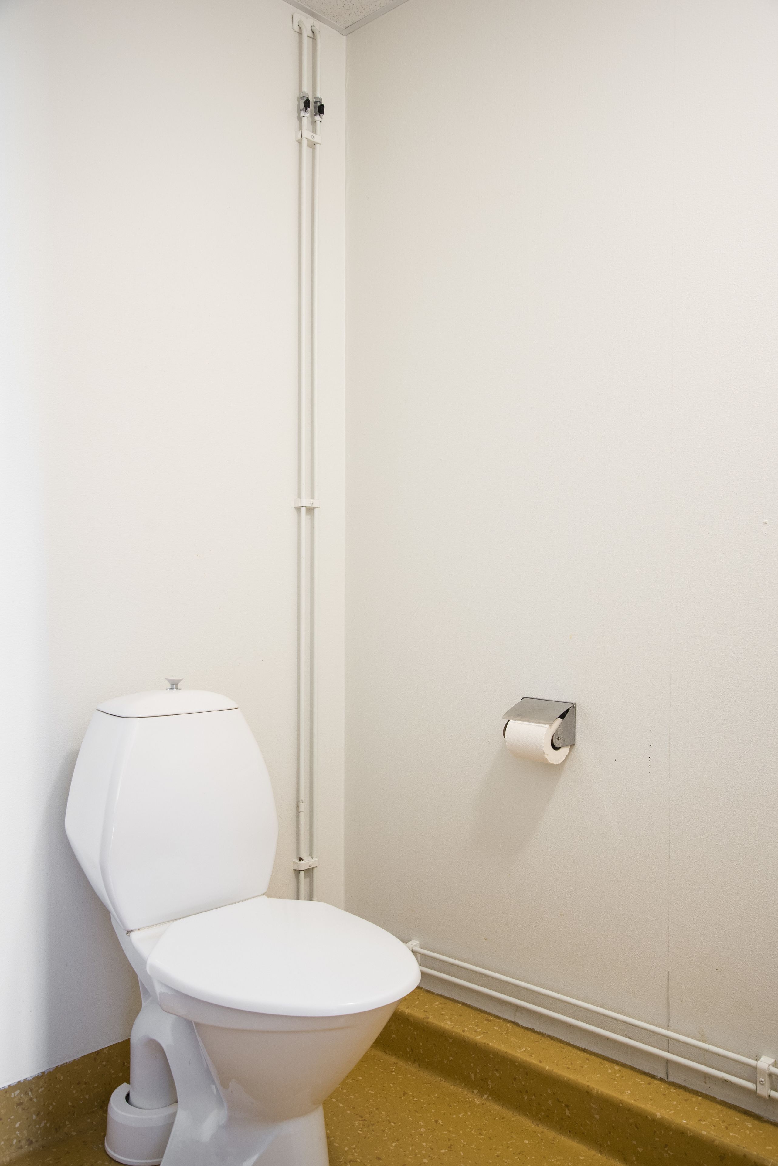 A white toilet with a flush handle, a white toilet paper holder, and a white pipe on the wall.