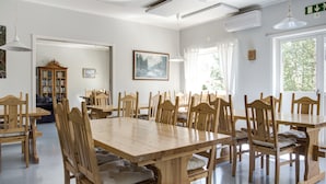 Een ruime eethoek met houten tafels en stoelen, een groot raam met gordijnen en een boekenkast op de achtergrond.