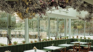 Un restaurante con un área de asientos con banco tapizado en verde, mesas blancas y plantas en macetas.