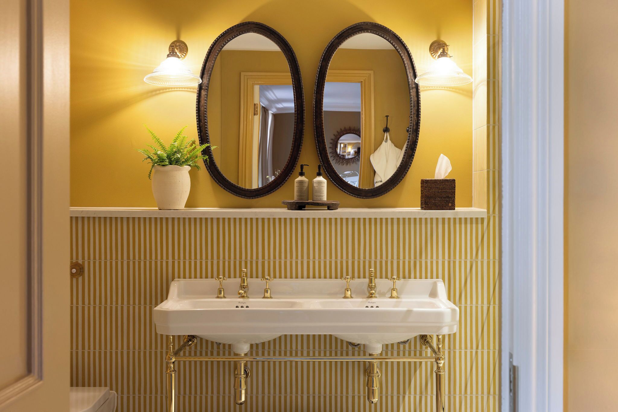 A bathroom with a double vanity, two wall-mounted mirrors, a potted plant, and a tissue dispenser.