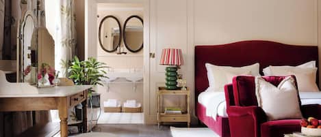 A bedroom with a large bed, a red armchair, a wooden desk, a mirror, and a bedside table with a lamp.